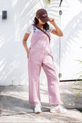 Woman wearing a pink overalls and brown cap standing outdoors with a white wall and palm tree in the background.