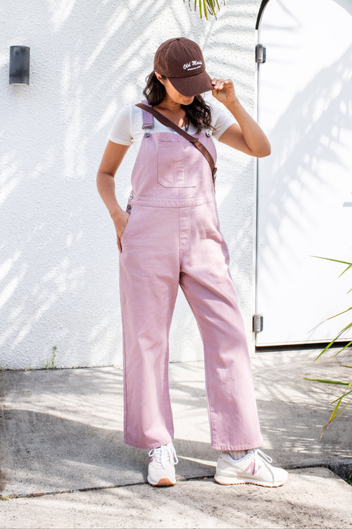 Woman wearing a pink overalls and brown cap standing outdoors with a white wall and palm tree in the background.
