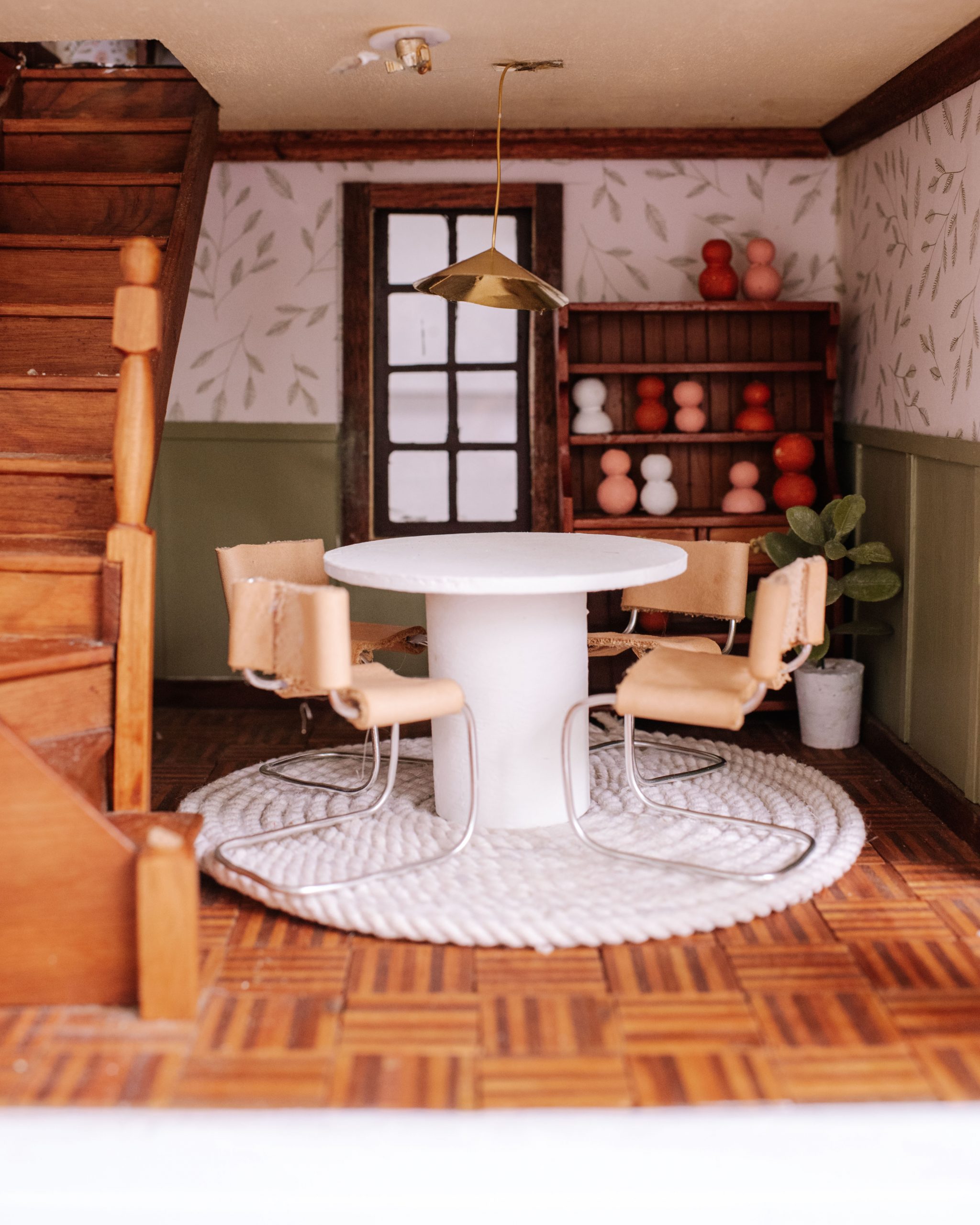 Image of a miniature dollhouse dining set. The table is a white round table made from a coaster and cardboard tube from a wrapping paper roll. The chairs have a metal frame and leather seats.
